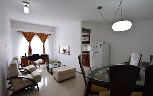 Open-plan living and dining space: round glass dining table with dark chairs, beige seating, and a kitchen with a white refrigerator in the background.