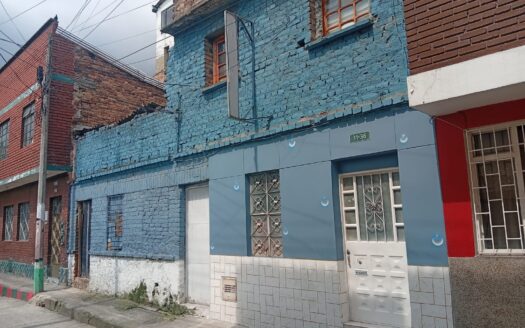 Blue-painted brick building on a street with metal window bars and tiled lower facade; neighboring red brick rowhouse on the left; utility wires overhead.