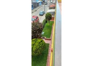 View from a balcony of a street with parked red and blue cars and a small green yard with trimmed shrubs and a tree below the railing.