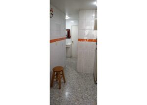 Narrow white-tiled hallway/kitchen with an orange tile border, a wooden stool in the foreground, and a sink visible toward the back left.