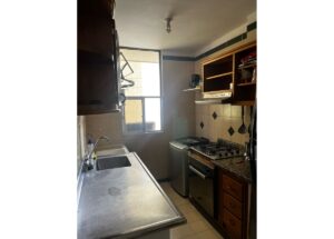 Small kitchen with a stainless steel sink and countertop, stove with oven, and a washing machine beside wooden shelves near a window.