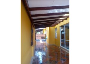 Covered outdoor walkway with yellow walls and dark wooden beams, and a wet glossy tiled floor leading to a wooden gate at the end.