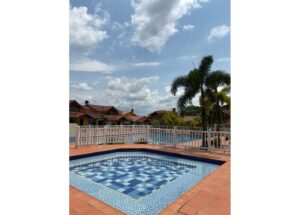Poolside view at a resort: blue mosaic pool with terracotta deck, white railing, and tall palm trees under a blue sky with clouds.