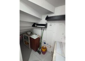 Small utility/laundry corner with a sink, two shelves, and cleaning supplies (broom, mop, bucket) against white walls and exposed piping.