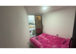 Small bedroom with pink patterned duvet on a bed near a balcony door showing city buildings outside.