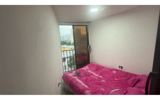 Small bedroom with pink patterned duvet on a bed near a balcony door showing city buildings outside.