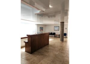 Wooden reception desk in a bright waiting area with blue chairs along the wall and framed artwork on the walls.