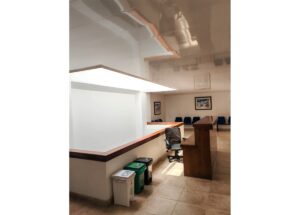 Wooden courtroom podium with a black swivel chair, row of blue chairs along the back wall, and waste/recycle bins nearby by a white partition.