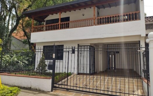 Two-story white house with a wooden balcony, black metal gate, tiled driveway, and greenery along the left.