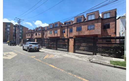Row of brick townhouses behind a tall black slatted fence, with a silver SUV parked on the street under a bright blue sky.