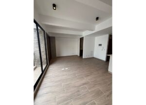 Empty modern living room with large sliding glass doors to a balcony, wood-look tile floor, white walls and ceiling beams.