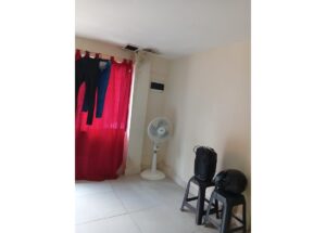 A dim room with a red sheer curtain, clothes hanging on a rod, and a standing fan near the wall. Two stools hold a backpack and a helmet; visible mold near the ceiling.