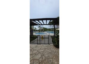 Gated view overlooking a resort pool area with a stone patio, black fence and wooden pergola surrounded by palm trees.
