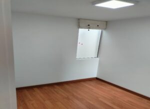 Small empty room with white walls and wood flooring, featuring a single window with a shade and a ceiling light above.