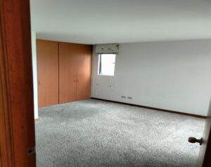 Empty bedroom with built-in wooden closet doors along one wall, a small window with a valance, and gray carpeted floor.