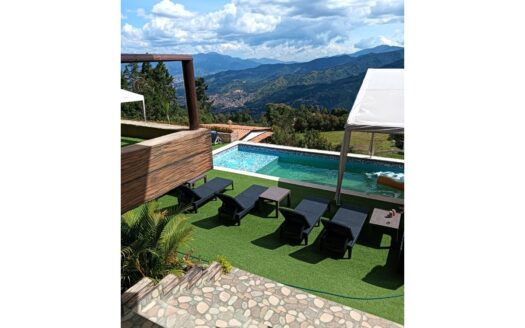 Poolside deck with black lounge chairs and small tables on green turf, stone steps in foreground, and mountain view under a partly cloudy sky.