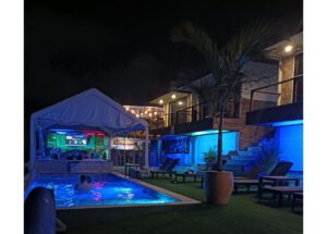 Nighttime poolside area with a shaded canopy, blue accent lighting, and lounge chairs along the pool.