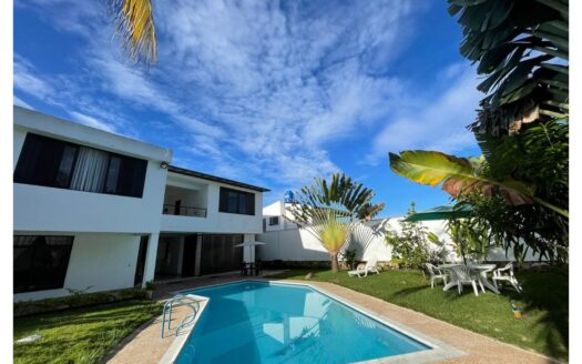 White two-story villa with a blue rectangular pool in a sunny yard, with patio furniture and palm trees nearby.
