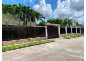 Wide shot of a gated residential property: a long black metal fence on a stone base, with a central gate, facing a sunny street and palm trees behind under a blue sky with clouds.