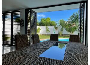 Wicker outdoor dining table and chairs on a covered patio, overlooking a backyard pool and tropical plants under a blue sky.