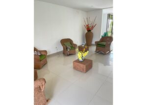 Wicker seating area with four rattan chairs and a square coffee table adorned with yellow flowers, in a bright lobby corner.