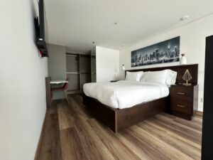 Bright hotel bedroom with a large white bed, dark wood headboard, and matching nightstands; cityscape artwork above the bed.
