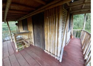 Bamboo hut porch with a dark wooden door, bamboo walls, and a red wooden deck surrounded by railing.