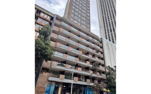 Tall brick apartment building with balconies along a busy city street, pedestrians and bicycles in front.