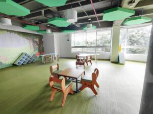 Bright children's playroom with small wooden animal-shaped chairs around a square table on a green striped carpet, large windows, and colorful ceiling panels. Alternative: A kid-friendly classroom area featuring animal-shaped chairs, low tables, a mural wall, and abundant natural light from big windows.