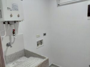 Utility laundry room with a wall-mounted white water heater, hoses, and a corner concrete utility sink with faucet fixtures.