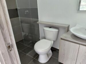 Modern bathroom with a white toilet under a floating shelf, a wooden vanity with a round vessel sink, and a glass-walled shower.