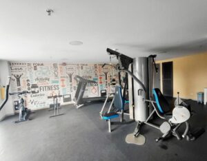 Gym with a large multi-station weight machine in the foreground and a wall with fitness-themed word art on the left wall.