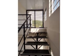 Industrial metal staircase in a bright stairwell with black railings and large windowed wall at the top corridor guard light.