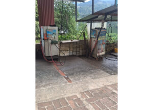 Two vintage gas pumps with red hoses at an outdoor station on a brick and concrete forecourt, hillside and trees visible in the background.