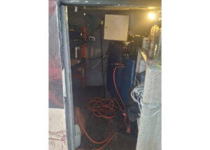 Cluttered workshop alcove featuring a blue cylindrical tank, orange extension cords on the floor, and a workbench with tools.