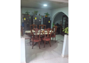 Round wooden dining table with eight carved chairs in a kitchen/dining area, refrigerators along the back wall and potted plants on top.