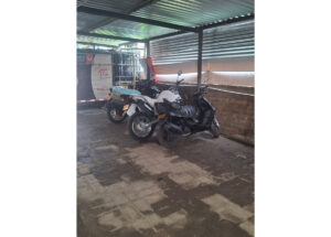 Three motorcycles parked under a metal canopy beside a brick wall, one Yamaha, with a delivery truck in the background.