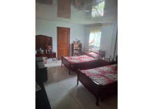 Twin bedroom with two wooden beds, a dresser with mirror, and a small bookshelf against light blue walls near a window with sheer curtains.