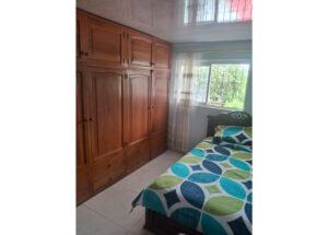 Bedroom with a tall wooden wardrobe along the left wall, a single bed with a teal and lime geometric quilt, and a window with sheer curtains.