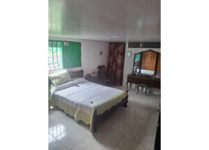 Bedroom with a large bed, pale green bedspread, and pillows; green window blinds and a floral curtain on the side. A wooden vanity with mirror and matching stool sits against the wall.