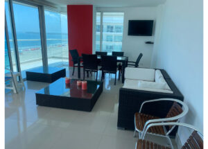 Modern living area with white tiled floor, black wicker sofa set, glass coffee table, and a wall-mounted TV in a high-rise apartment overlooking the ocean.