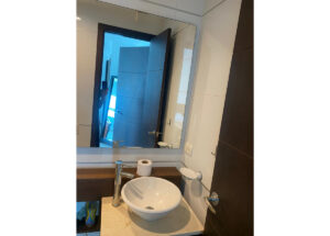 Bathroom vanity with a round white vessel sink, chrome faucet, and a roll of toilet paper on a beige countertop; open door reflects in the mirror beyond the sink.