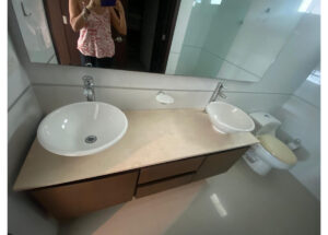 Bathroom vanity with two round vessel sinks, beige countertop, and a toilet to the right; person taking the photo is visible in the mirror.