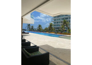 Outdoor hotel pool area with wicker lounge chairs, palm trees, and a modern building in the background on a sunny day.
