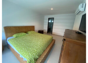 Bedroom with a wicker-platform bed, green geometric quilt, two pillows, a wooden dresser with a TV, and an overhead AC unit.