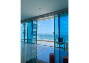 Indoor living room with glass doors opening to a balcony and ocean view; a paraglider in blue sky beyond the coast.