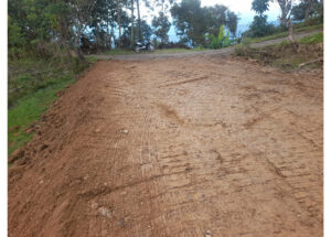 Unpaved dirt road under construction in a rural area, with a motorcycle parked at the edge and trees in the background.