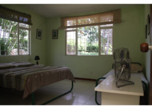 Bedroom with green walls, a bed on the left, a small bedside lamp, and framed photos on the wall; a large window reveals a garden outside.