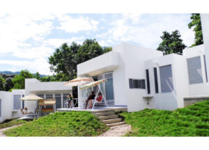 Modern white villa with an outdoor patio where four people sit under striped umbrellas on a grassy slope.