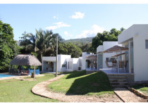 White modern villas with outdoor seating under striped umbrellas, palm trees, and a thatched gazebo by a pool in a tropical resort courtyard.